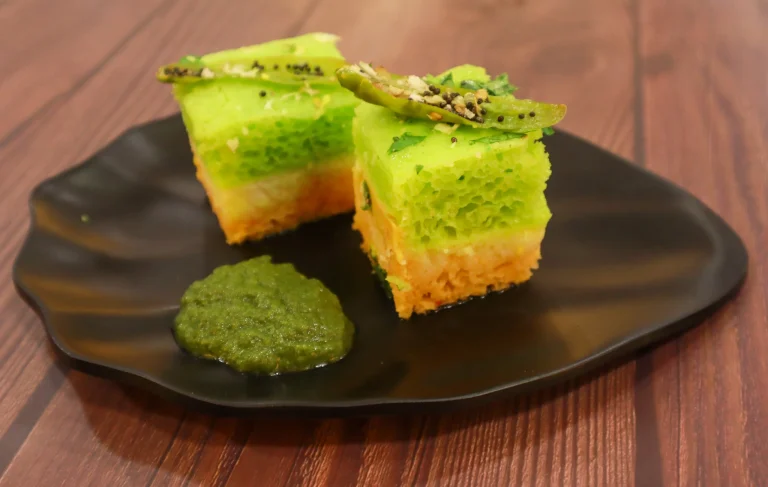 Gujarati dish served with a vibrant green and orange layered dhokla, garnished with chili and cilantro, accompanied by green chutney