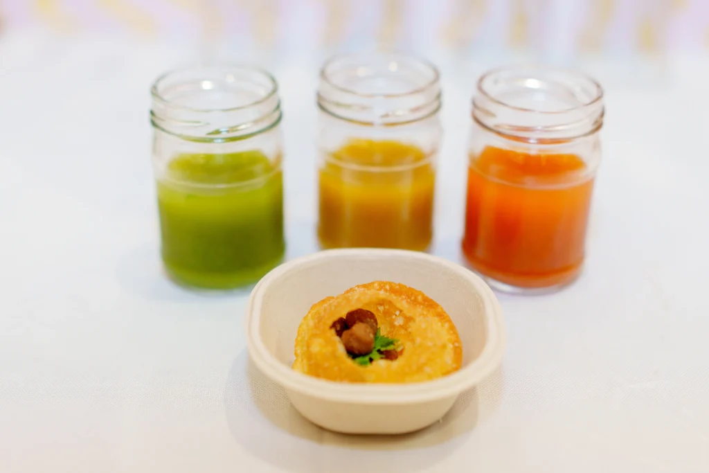 Pani puri served with three jars of flavored water (green, yellow, and orange) and garnished with coriander, served in a small container