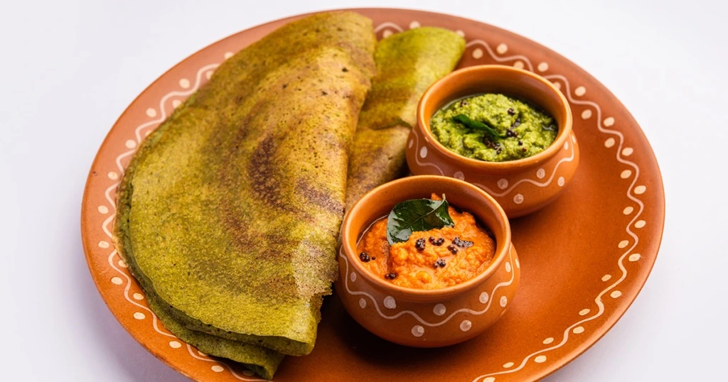 Traditional Andhra Pesaratttu (green dosa) served with chutneys on a terracotta plate